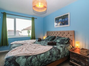 A bedroom with a bed, a window with curtains, a wall-mounted picture, and a bedside table with a lamp. - Home by the sea (Llandudno)