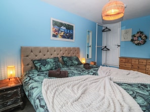Ein Schlafzimmer mit einer gepolsterten Kopfteile, einem Bett mit gemusterten Betttextilien, einer weißen Tagesdecke, einer Pendelleuchte, einem gerahmten Bild an der Wand und einer Holzkommode.