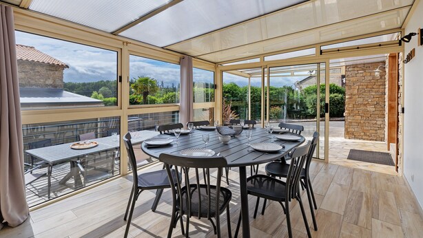 A covered outdoor dining area with a table and chairs, large windows, and a view of a stone building. - Le Clos des Prés with Pool, Spa and Table Football (Rives de l'Yon)