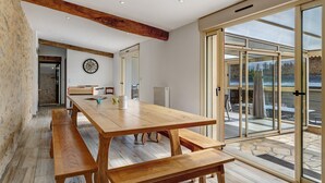 A spacious dining area with a wooden table and benches, large sliding glass doors leading to an outdoor patio, and a visible kitchen area in the background. - Le Clos des Prés with Pool, Spa and Table Football (Rives de l'Yon)
