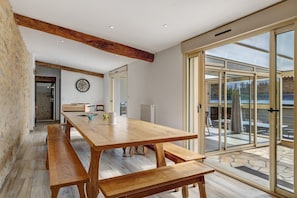 A spacious dining area with a wooden table and benches, large sliding glass doors leading to an outdoor patio, and a visible kitchen area in the background. - Le Clos des Prés with Pool, Spa and Table Football (Rives de l'Yon)
