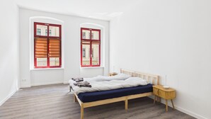 A bedroom with a wooden bed, two windows with red frames, a bedside table, and a nightstand. - Family apartment with balcony, up to 5 guests - GÖ04 (Görlitz)