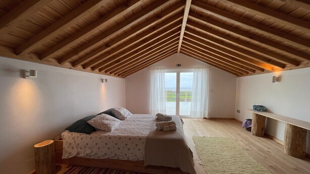 A bedroom with a sloped wooden ceiling, a bed with pillows and a blanket, a small wooden stool, and a window with a view. - Holiday Home 'Casa Baía Da Lagoa' with Sea View and Wi-Fi (Lagoa)