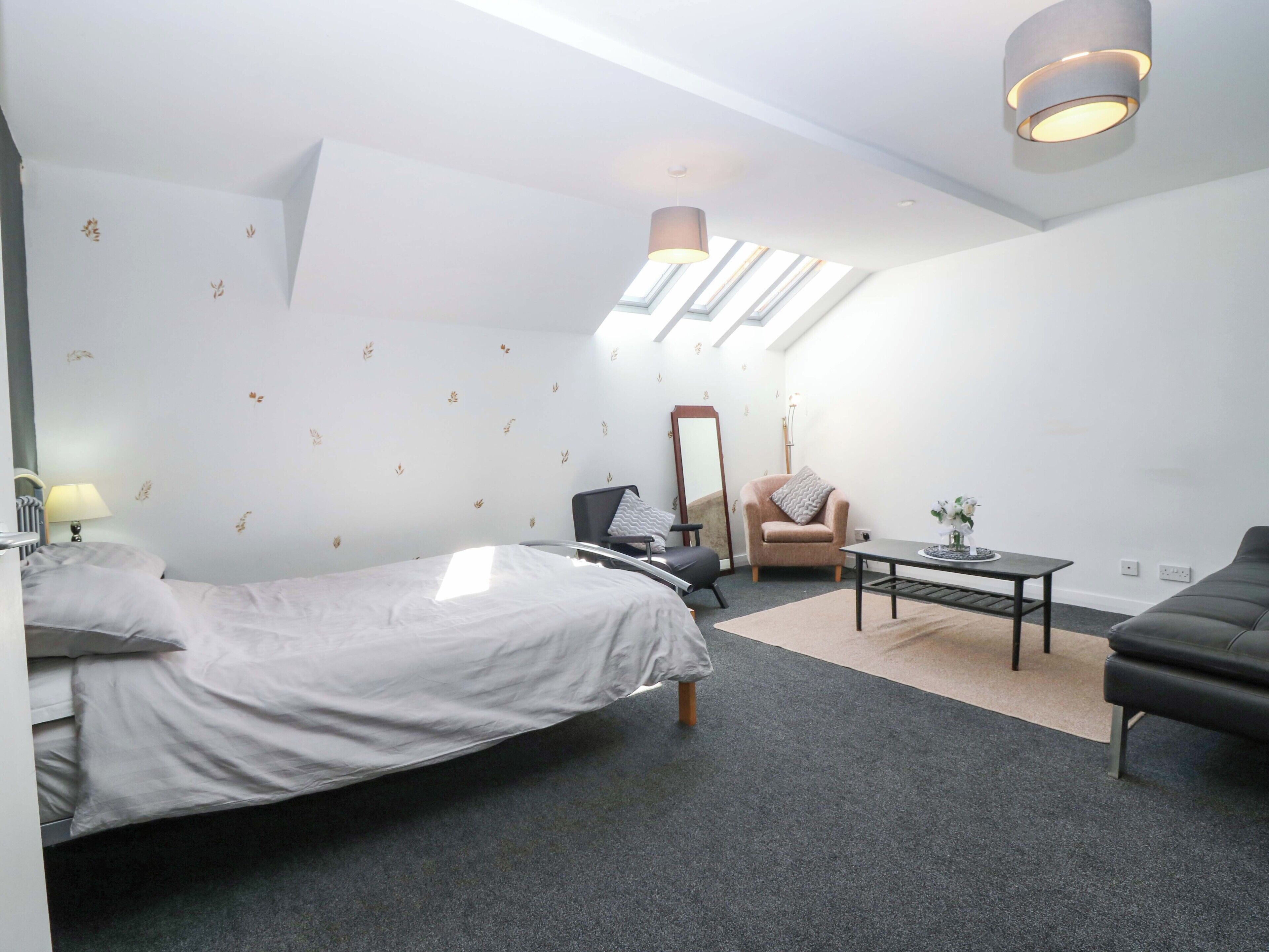 A modern hotel room with a bed, a chair, a table, and a sofa. There is a skylight and a mirror on the wall.