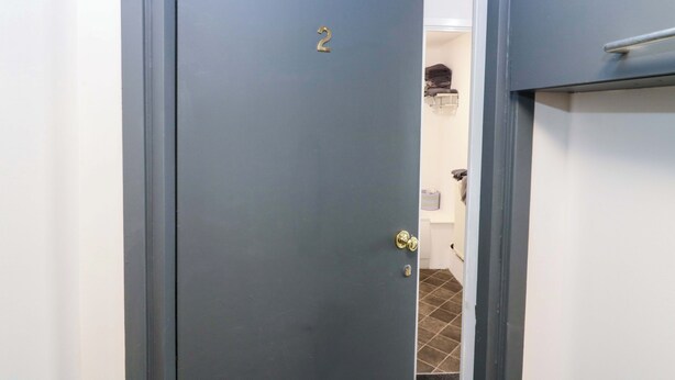 Eine geschlossene Tür mit der Zahl 2, einem Metallbeschlag und einem teilweisen Blick auf einen gefliesten Boden sowie eine weiße Wand.