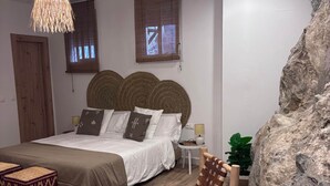 A bedroom with a bed, a woven chair, a small table, a potted plant, and a large rock feature.