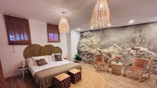 A bedroom with a bed, two woven poufs, a large woven rug, a rock wall, and two wicker chairs.