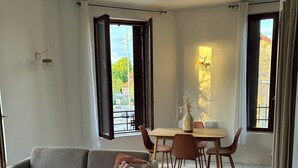 Un salon avec un canapé, une table à manger et des chaises, un vase rempli de fleurs et une vue sur l’extérieur par la fenêtre ouverte.