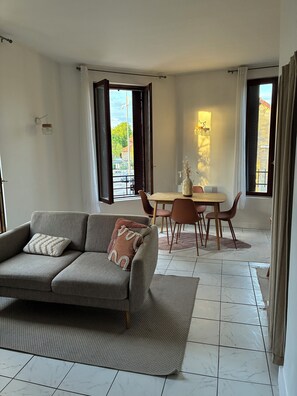 Ein Wohnzimmer mit einer Couch, einem Esstisch mit Stühlen, einer Vase mit Blumen und Blick durch das offene Fenster.