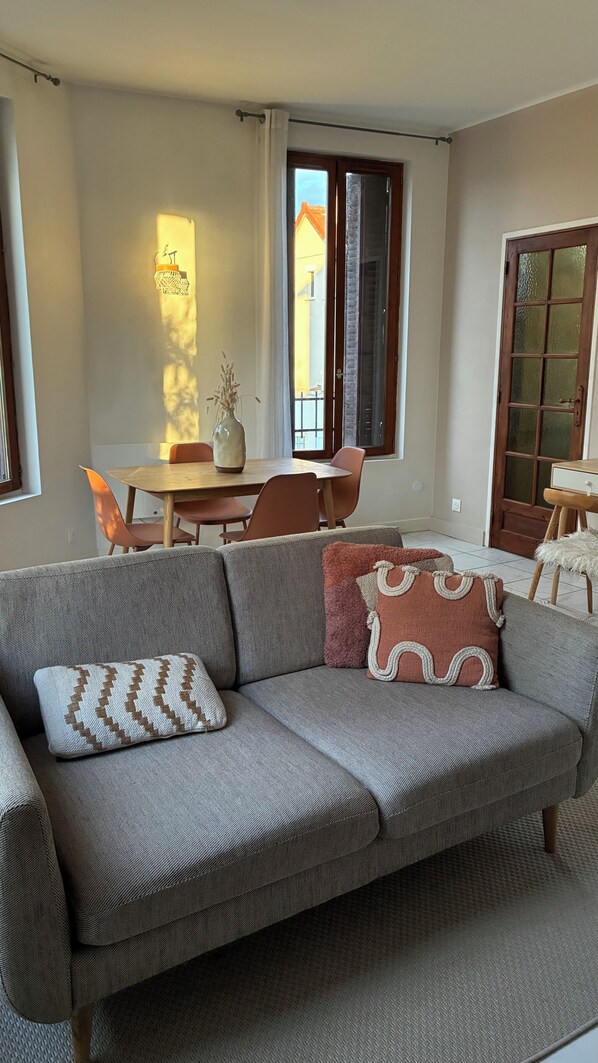 Ein Wohnzimmer mit einer Couch, einem Esstisch mit Stühlen, einem Fenster mit Vorhängen und einer Tür.
