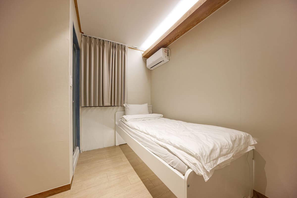 Quarto pequeno com cama de solteiro, teto inclinado, janela com cortinas e ar condicionado fixado na parede.
