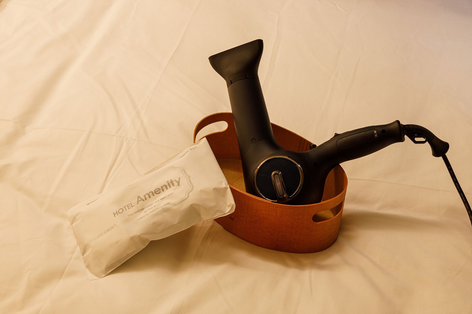 A black hairdryer and a white bag with 'HOTEL AMENITY' text in a brown basket on a bed.