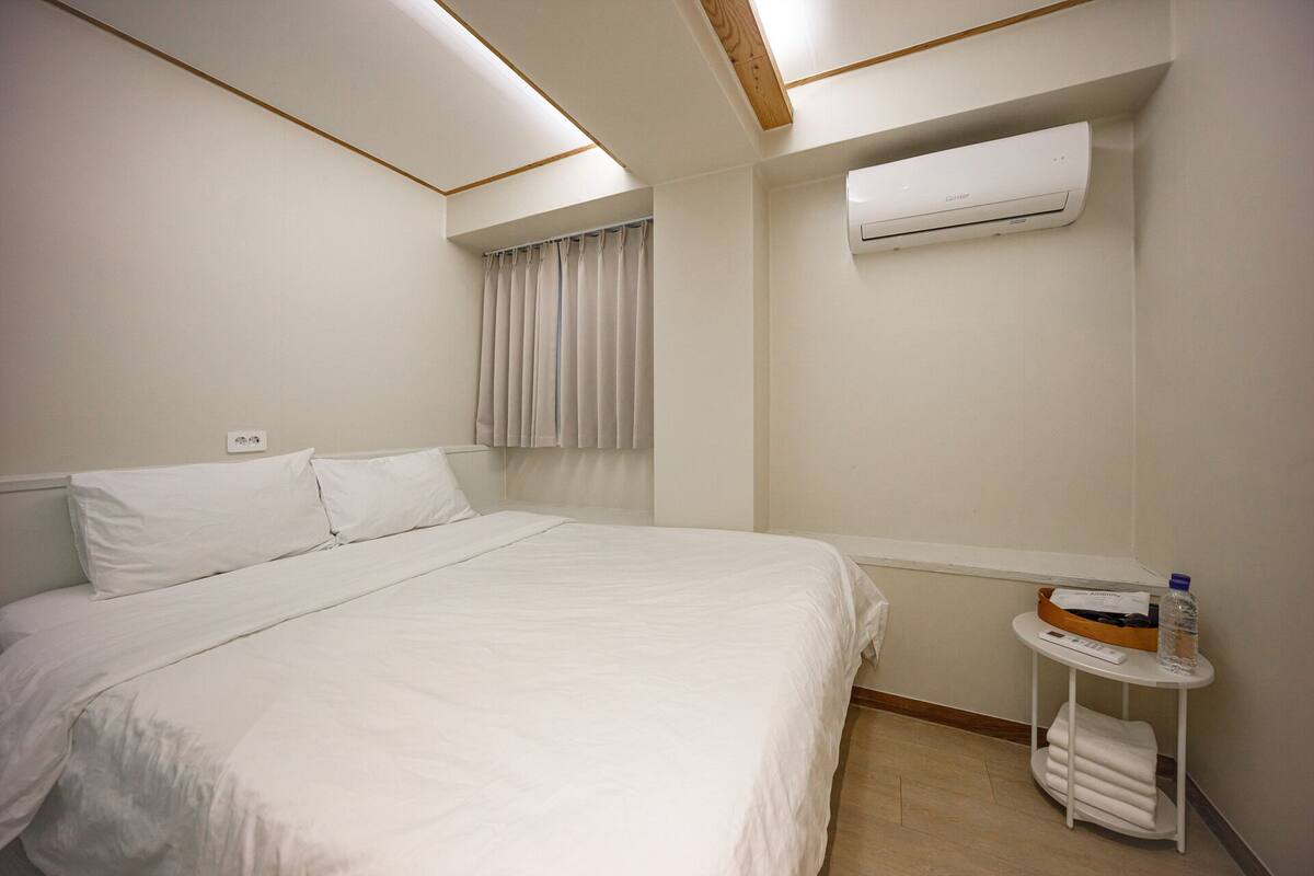 Quarto de hotel com cama, ar condicionado, janela com cortinas e uma pequena mesa com toalhas e artigos de higiene pessoal.