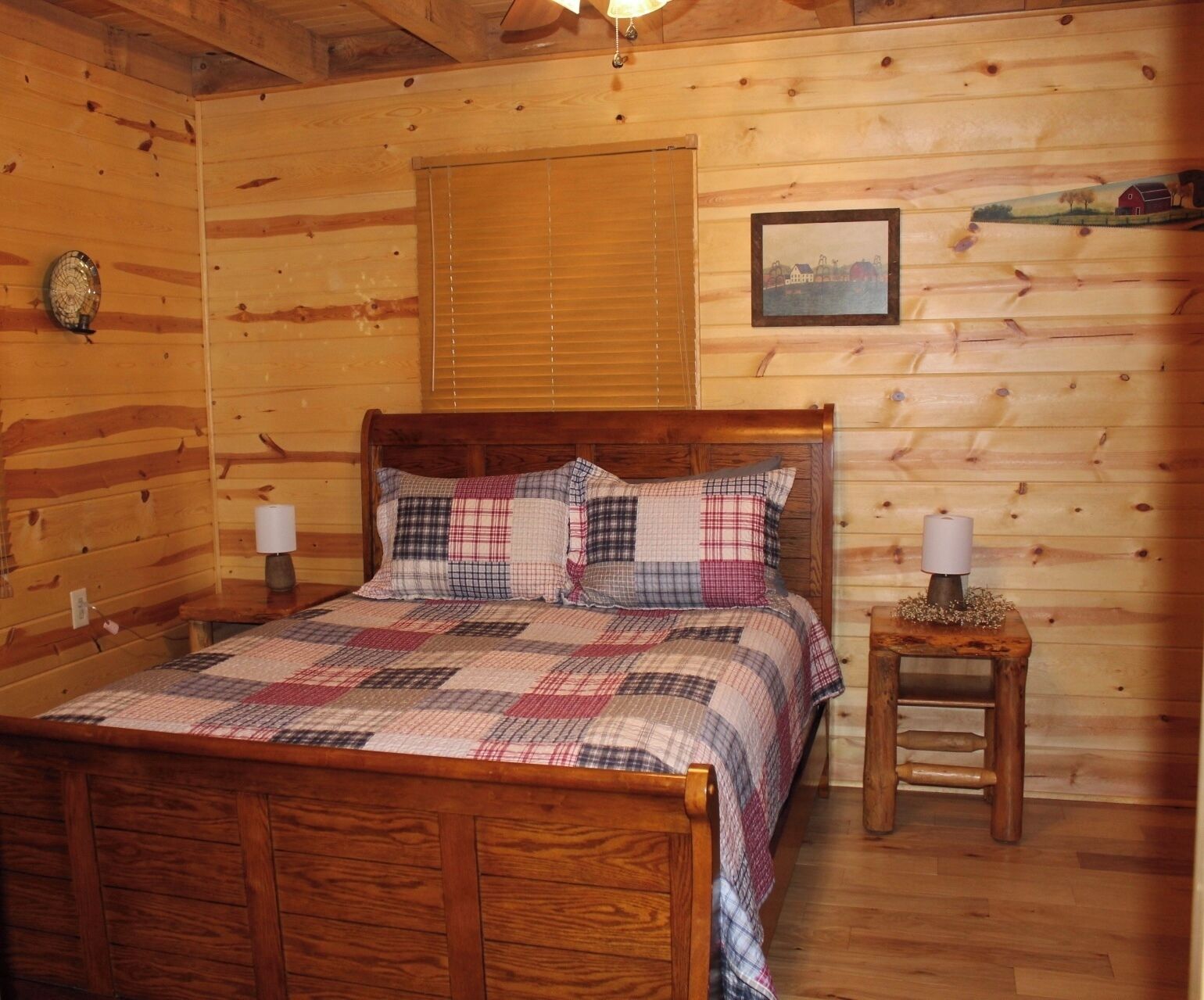 Ein Schlafzimmer einer Holzhütte mit einem Bett, zwei Nachttischlampen, einem kleinen Tisch mit Lampe, einem Fenster mit Jalousien und einem gerahmten Bild an der Wand.