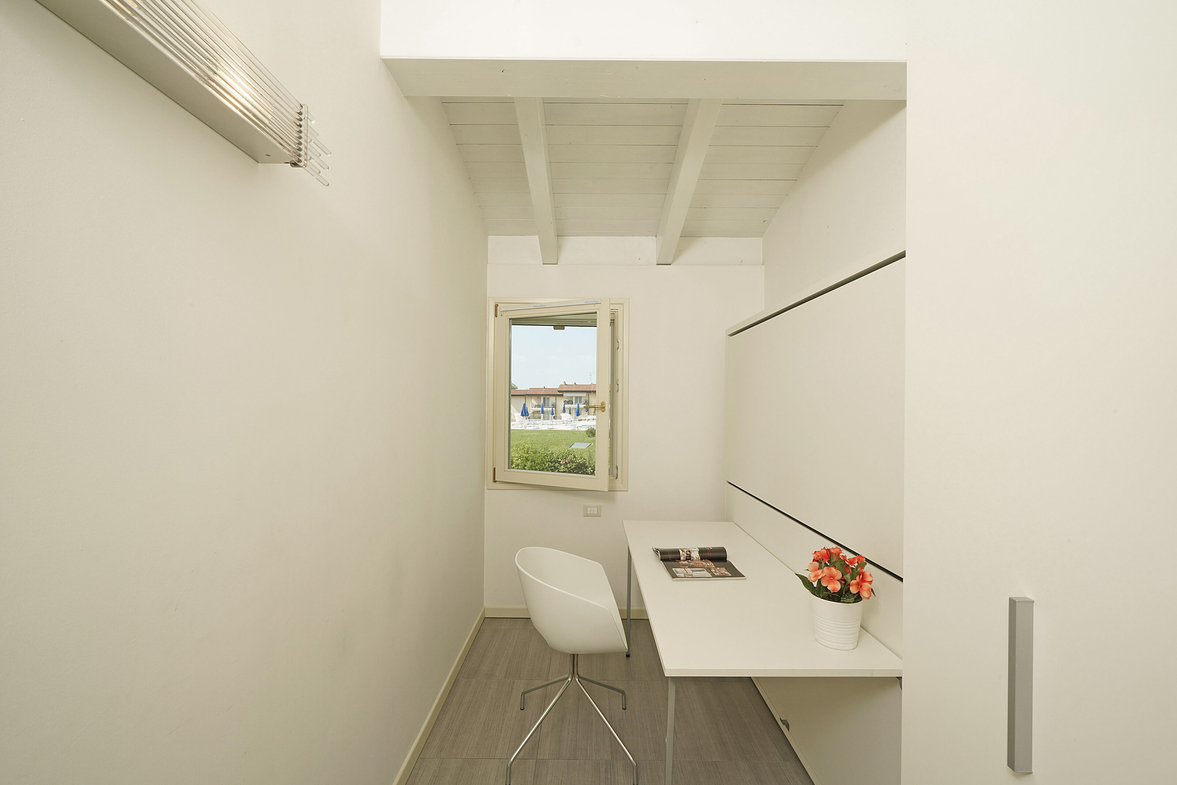 Une petite pièce moderne, dotée d’un bureau blanc, d’une chaise blanche, d’une fenêtre offrant une vue et d’une bouche d’aération au plafond.