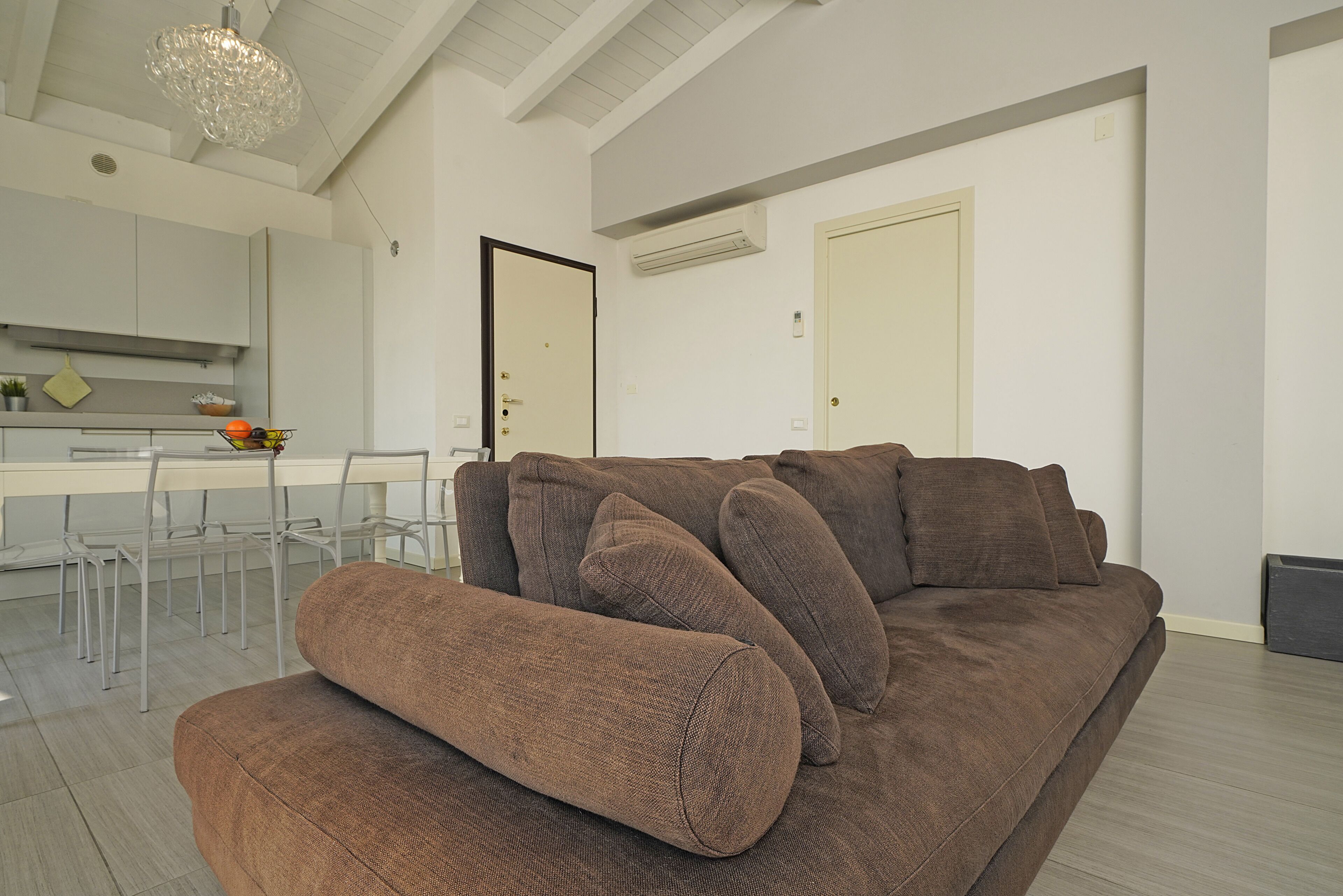 En moderne stue med en brun sofa, et spiseområde med et hvidt bord og stole, og et køkken i baggrunden.