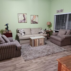 Ein Wohnzimmer mit Sofa, Sessel, Couchtisch und Teppich. Zwei Wandbilder und ein Fenster mit Jalousien sind zu sehen.