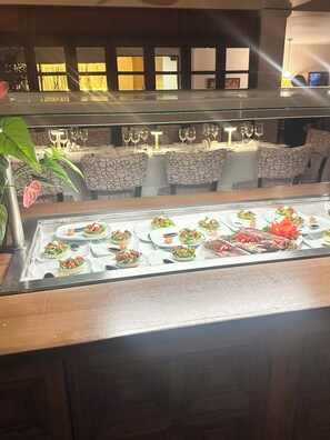 Restaurante com vitrine de vidro exibindo diversos pratos, uma planta e área de refeições com cadeiras e mesas estampadas.