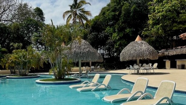 A swimming pool with lounge chairs and thatched umbrellas, surrounded by lush greenery and palm trees. - Coronado Luxury Club & Suites - 1 bedroom- Green fees for 2 & resort access (Panamá Oeste Province)