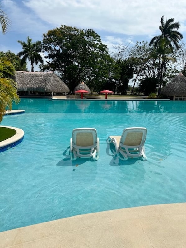 A swimming pool with lounge chairs, palm trees, and thatched-roof structures.