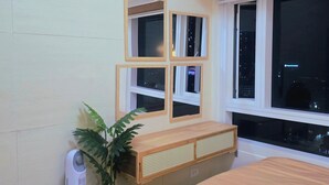 A bedroom with a bed, a bedside table, a plant, a radiator, and a window with a city view. - Dyad Suites Pasay (Pasay)