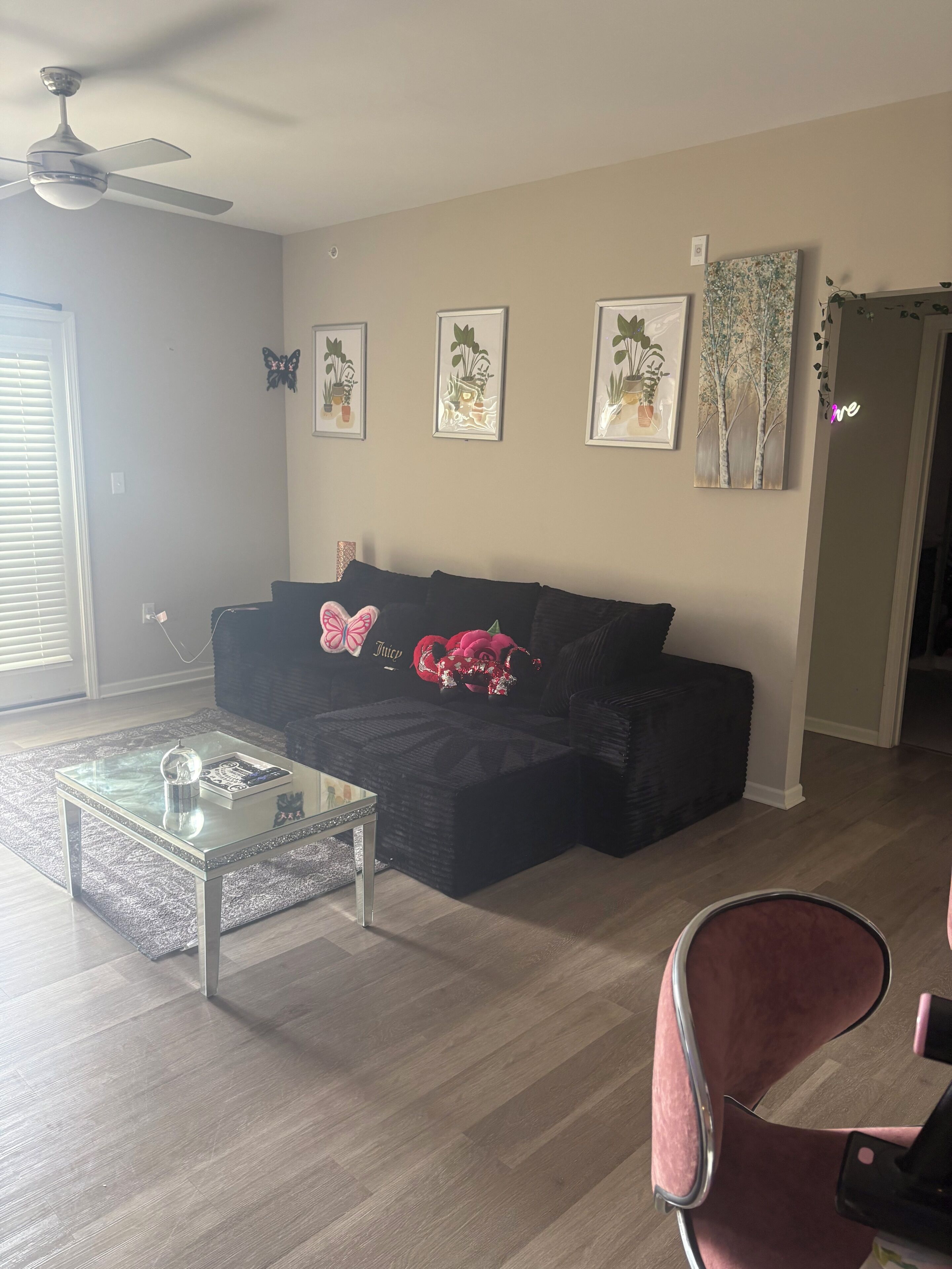 Een woonkamer met een glazen salontafel, een zwarte bank met decoratieve kussens, ingelijste botanische prints aan de muur en een plafondventilator.