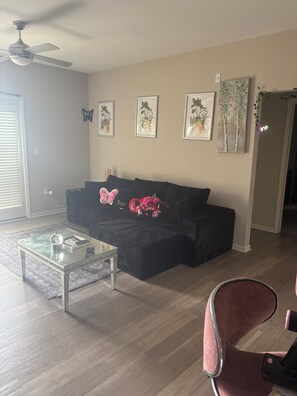 A living room with a glass coffee table, a black sofa with decorative pillows, framed botanical prints on the wall, and a ceiling fan.