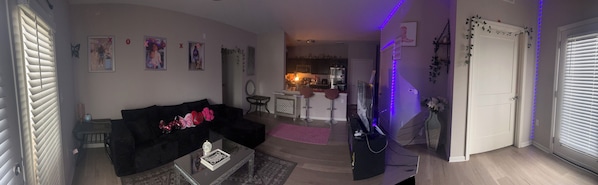 A living room with a black sofa, a coffee table, and a TV. There is a kitchen in the background with a refrigerator and a dining area.