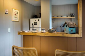 Eine Küchenzeile mit einem weißen Kühlschrank, einer Kaffeemaschine, einem kleinen Tisch mit Schüssel und Tassen sowie einem blauen Vogelhaus auf der Arbeitsplatte.
