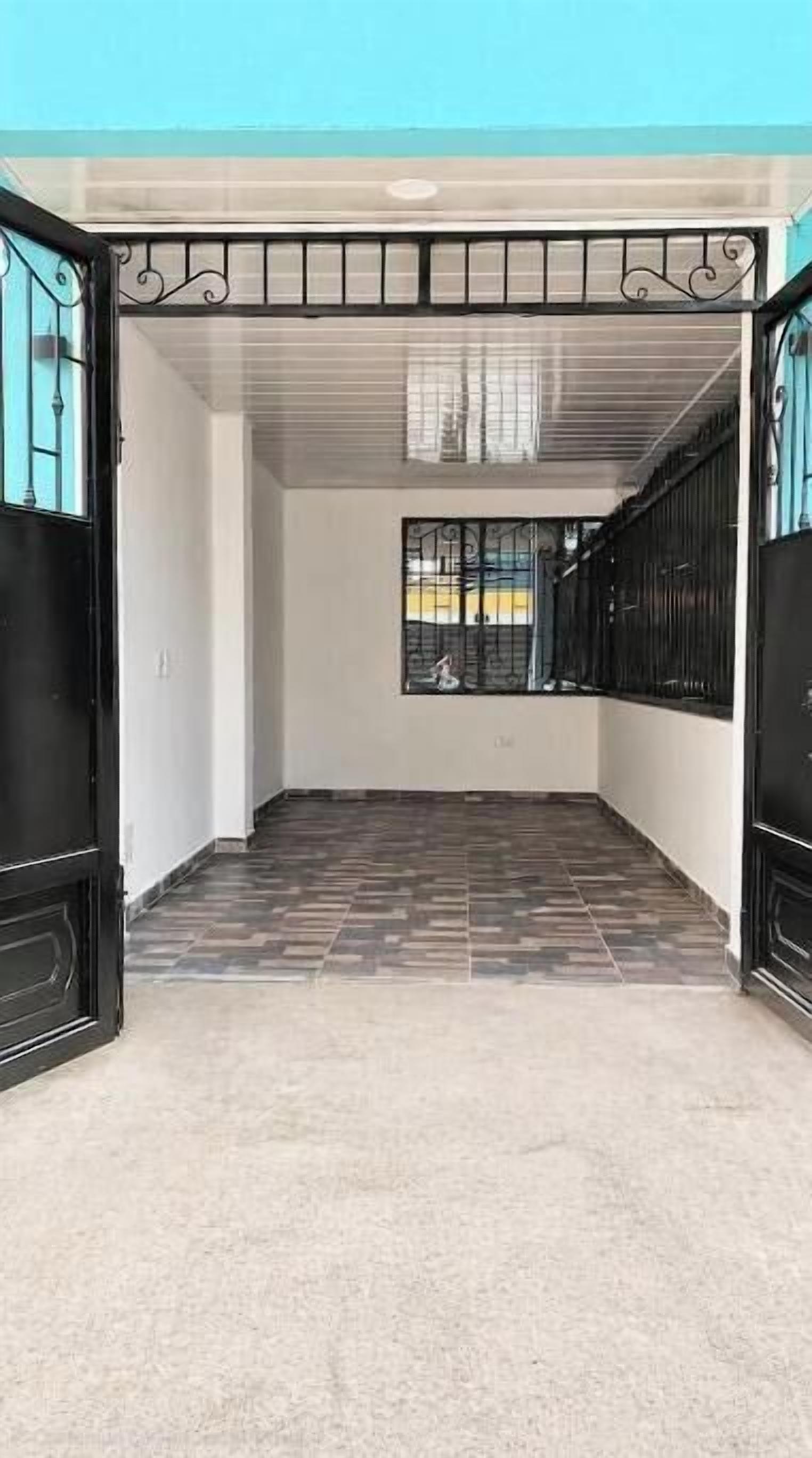 Un pasillo vacío con suelo de baldosas, una ventana con barrotes y un portón negro.