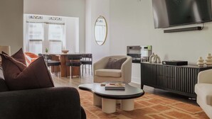 Ein modernes Wohnzimmer mit Sofa, Sessel, Couchtisch, Fernseher und einem Essbereich mit Stühlen und Tisch.