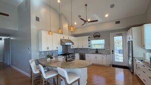 Een moderne keuken met witte kasten, een centraal eiland met barkrukken, hangende verlichting, een plafondventilator en uitzicht buiten.