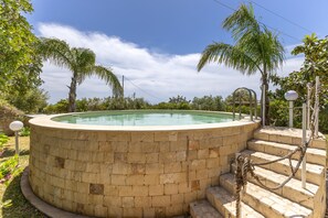 Une piscine circulaire en pierre, accessible par des marches, entourée de verdure et surplombée d’un ciel dégagé.