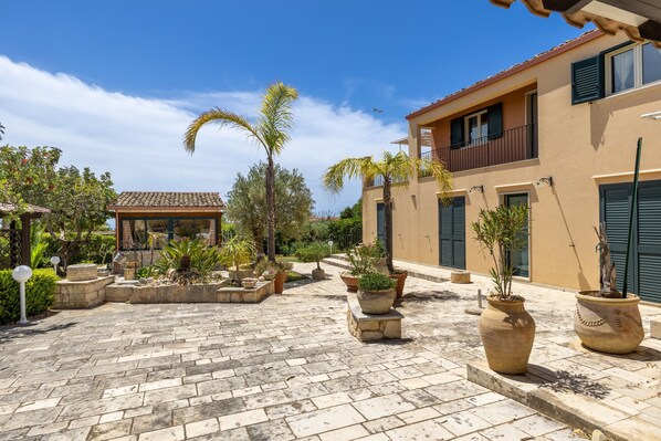 A courtyard with stone paving, potted plants, a palm tree, and a building with balconies and shutters.