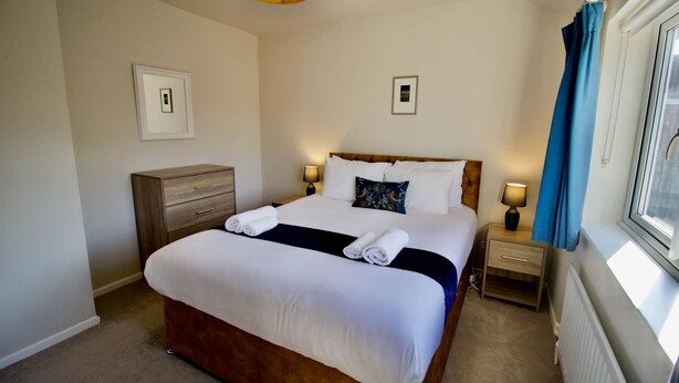 Een net opgemaakt bed met witte lakens en een donker accent, een houten nachtkastje met een lamp, en een raam met blauwe gordijnen.