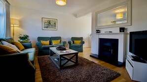 Een woonkamer met een open haard, een televisie en twee groene fauteuils met gele kussens.