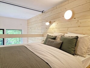 Ein Schlafzimmer mit einem Holzkopfende, einem Bett mit vielen Kissen, einem Fenster mit Blick auf Bäume und einer runden Leuchte.
