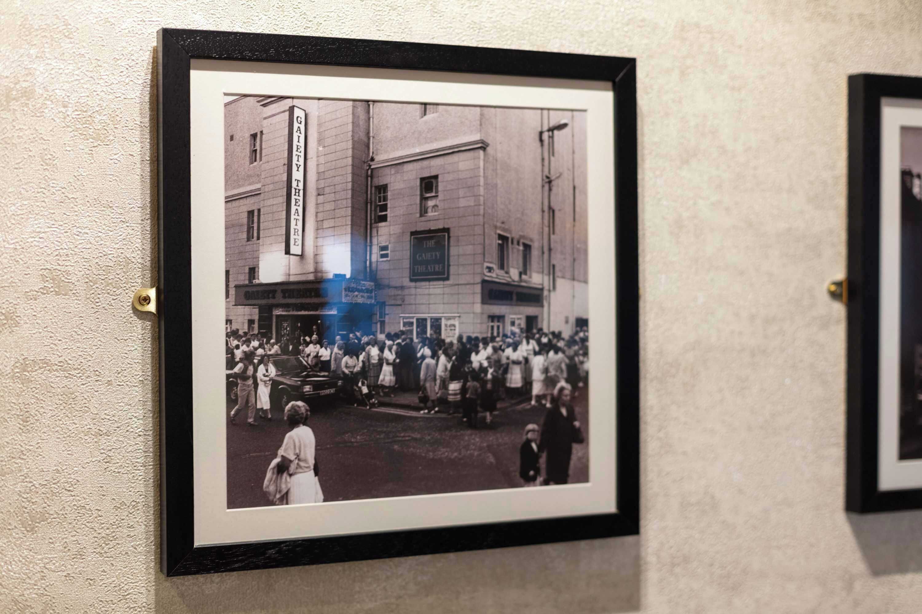 Ein gerahmtes Schwarz-Weiß-Foto einer belebten Straßenszene mit einem Gebäude mit der Aufschrift 'The Gaiety Theatre'.