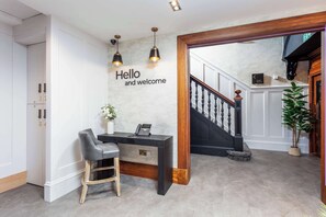 Un comptoir d’accueil avec une pancarte indiquant « Bonjour et bienvenue », un téléphone et une chaise. On aperçoit, à l’arrière-plan, un escalier avec une rampe en bois et un pot de fleurs.