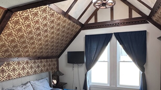 A bedroom with a sloped ceiling, a bed with a plaid bedspread, a blue throw blanket, a television, and a window with curtains. - Elegant Studio Near Boston (Malden)