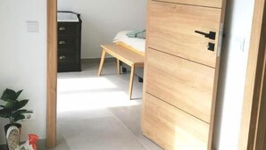 Una habitación con puerta de madera, una cama con manta azul, una mesita y un aparador.