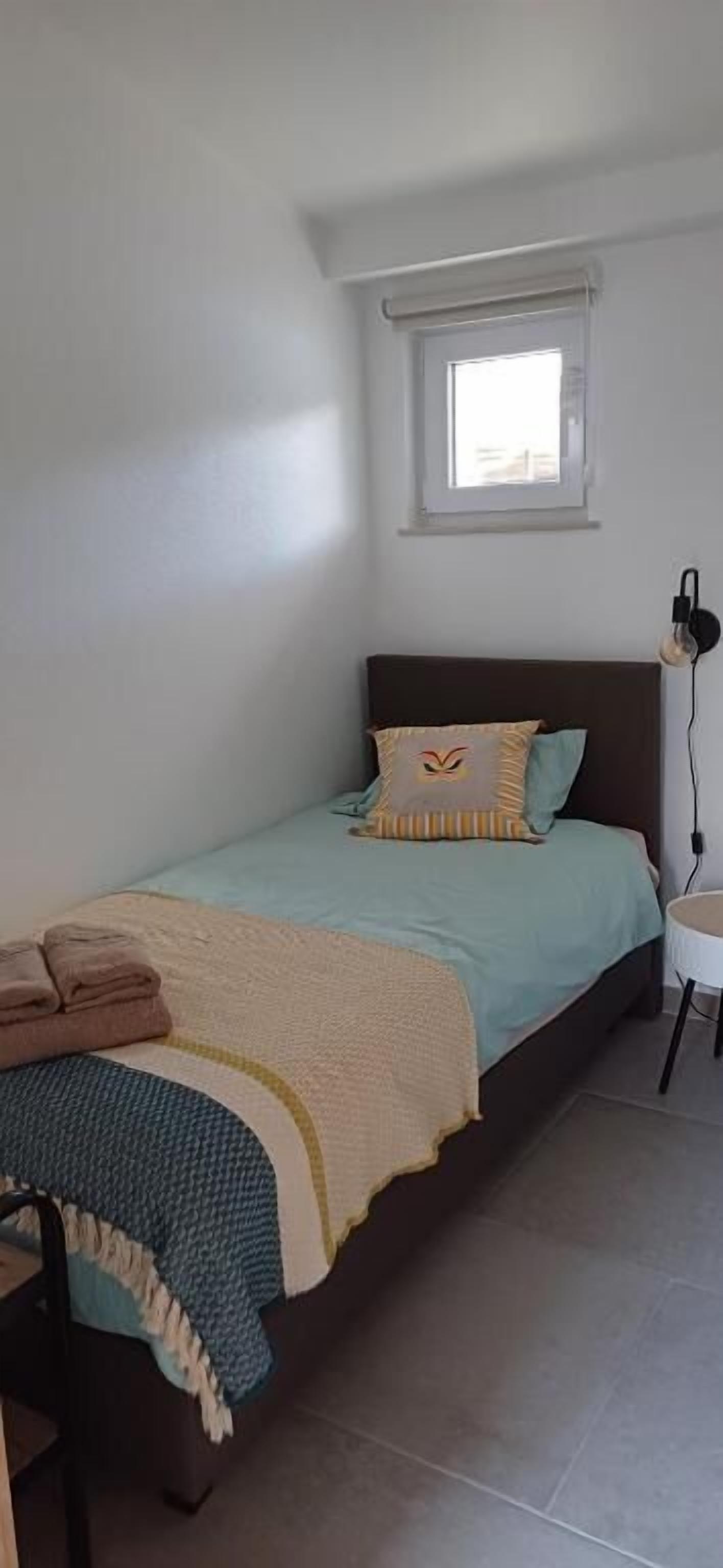 Una cama individual con cabecera, una mesita de noche con lámpara y una ventana sobre la cama.
