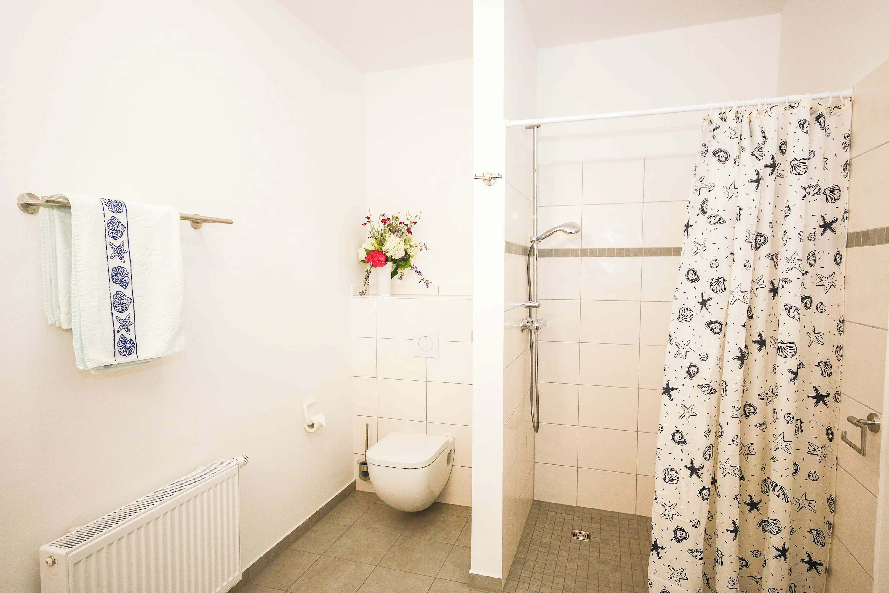 A bathroom with a toilet, a shower, a towel rack with a towel, and a flower vase on the wall.
