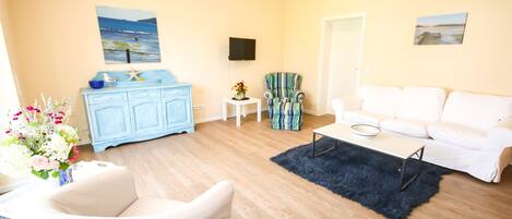 A living room with a white sofa, a blue cabinet, a small table with a vase, a blue rug, and a television on the wall.