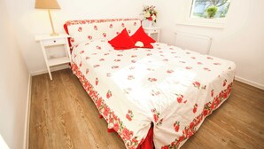 A bedroom with a floral bedspread, a nightstand with a lamp, a dresser, and a window with a view of trees. - FeWo Cottage im Ferienidyll am Großen Wentowsee (Gransee)
