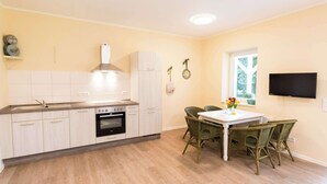 A kitchen with white cabinets, a built-in oven, and a dining area with wicker chairs and a white table. - FeWo Cottage im Ferienidyll am Großen Wentowsee (Gransee)