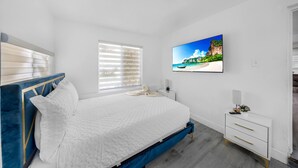A bedroom with a bed, a TV on the wall, a bedside table, and a window with blinds. - Miami Tree House Renovated Beach Jacuzzi (Miami)