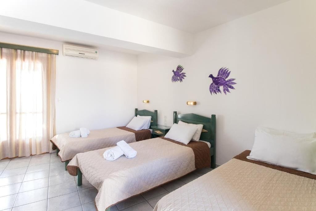 A hotel room with three single beds, a window with curtains, an air conditioning unit, and wall-mounted light fixtures.