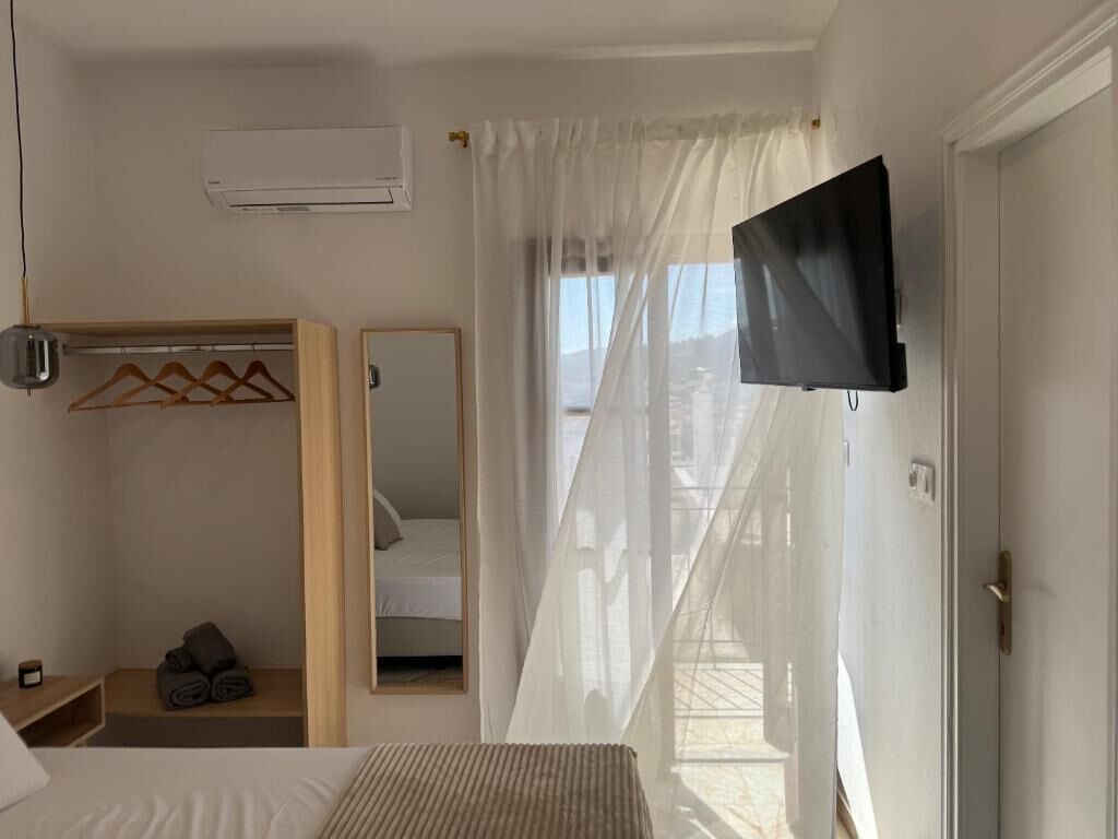 A hotel room with a bed, a wardrobe, a mirror, a flat-screen TV, and a window with sheer curtains.