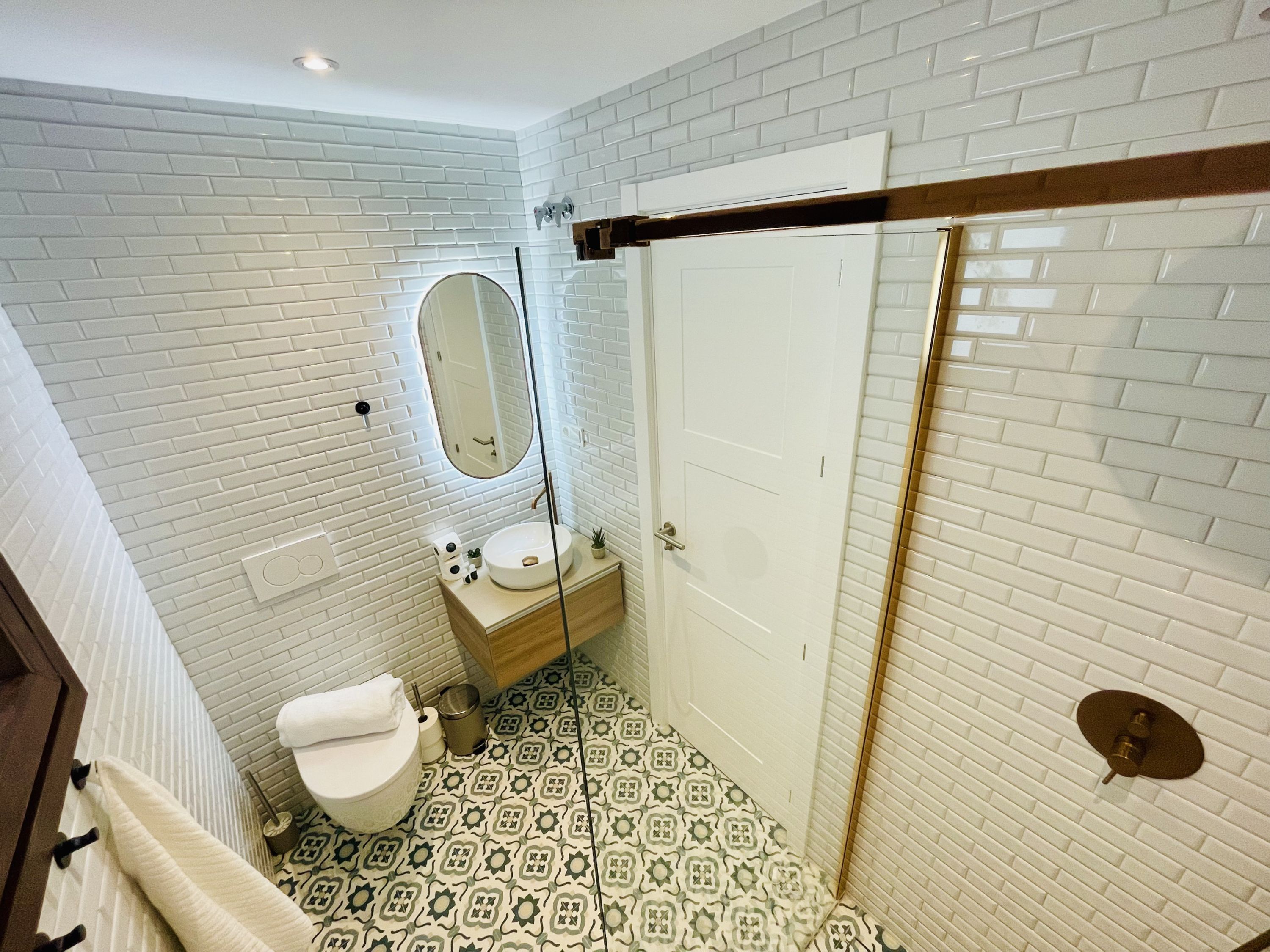 A bathroom with white brick walls, a white toilet, a sink with a mirror, patterned floor tiles, and a door with a handle.